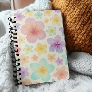 Puede incluir: Un cuaderno con espiral con una cubierta crema adornada con flores de acuarela en tonos pastel de rosa, morado, amarillo y azul. El cuaderno está sobre una superficie blanca texturizada.