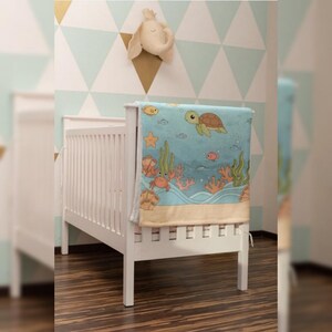 Puede incluir: Una cuna blanca con una manta azul con una escena submarina con una tortuga, peces y coral. La manta está sobre el lado de la cuna. Una cabeza de elefante de peluche decora la pared sobre la cuna. El fondo tiene un patrón geométrico.