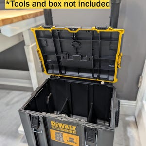 May include: Black and yellow DEWALT TOUGH SYSTEM 2.0 rolling tool box with an open lid. The box has a handle and wheels for easy transport. The text "Tools and box not included" is visible.