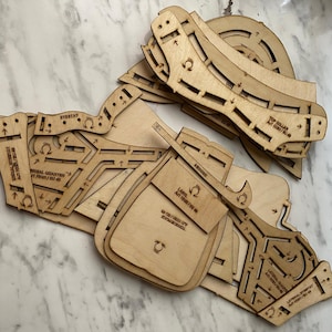 Puede incluir: Un conjunto de piezas de madera cortadas con láser, probablemente para un modelo o proyecto de manualidades. Las piezas tienen varias formas y tamaños, con marcas y texto grabados, incluyendo "EYESTAY" y "TOP DOLLAR". La madera es de color claro natural.