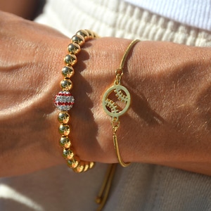 May include: Two gold bracelets with baseball charms. One bracelet has a beaded chain with a baseball charm with red and white stripes and clear rhinestones. The other bracelet has a thin chain with a baseball charm.