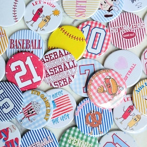 May include: A collection of circular pin-back buttons featuring various sports and food-themed designs, including baseballs, softballs, tennis, popcorn, hot dogs, pretzels, and text such as Baseball, Softball, Stormers, and The way the truth the life.