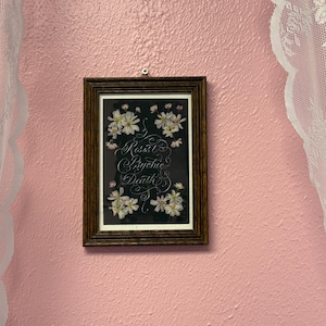 May include: A framed artwork with a dark background, adorned with white flowers and elegant script that reads "Rosal Psychic Death." The frame is a dark wood color, and the artwork is hanging on a textured pink wall, with white lace curtains on either side.