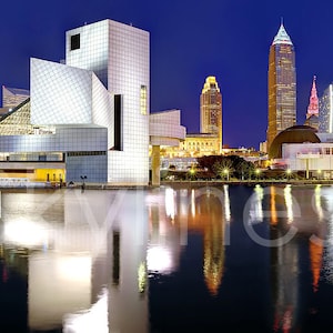May include: Panoramic view of the Cleveland, Ohio skyline at dusk, reflecting in the water. The image features iconic buildings, including the Rock & Roll Hall of Fame, and a stadium, under a deep blue sky. The word "Cleveland" is written in cursive.