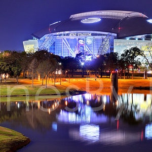 May include: A panoramic night view of a large, modern stadium with a reflective pond in the foreground. The stadium is illuminated with bright lights, creating a colorful reflection in the water. Trees line the edge of the pond and the stadium.