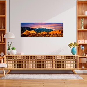 Puede incluir: Una impresi&oacute;n panor&aacute;mica en lienzo de una cordillera al atardecer. La obra presenta picos nevados, formaciones rocosas doradas y un cielo vibrante con tonos de naranja, rosa y azul. La impresi&oacute;n se muestra sobre un mueble de madera.