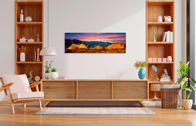 Puede incluir: Una impresi&oacute;n panor&aacute;mica en lienzo de una cordillera al atardecer. La obra presenta picos nevados, formaciones rocosas doradas y un cielo vibrante con tonos de naranja, rosa y azul. La impresi&oacute;n se muestra sobre un mueble de madera.