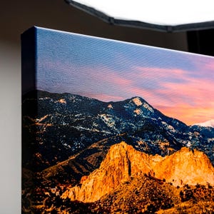 Puede incluir: Impresi&oacute;n en lienzo de un paisaje monta&ntilde;oso al atardecer. La imagen muestra formaciones rocosas doradas en primer plano, con monta&ntilde;as oscuras y nevadas y un cielo rosa y azul vibrante en el fondo.