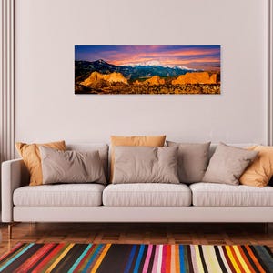 Puede incluir: Una impresi&oacute;n panor&aacute;mica en lienzo de una cordillera al atardecer. El cielo transita del azul profundo al naranja y rosa vibrantes, con picos nevados y terreno rocoso en primer plano. La obra de arte se exhibe sobre un sof&aacute; blanco.