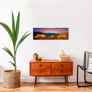 Puede incluir: Una impresi&oacute;n panor&aacute;mica de un paisaje monta&ntilde;oso con picos nevados bajo un cielo vibrante al atardecer. La obra de arte se exhibe sobre una c&oacute;moda de madera de estilo moderno, con una planta en maceta y objetos decorativos.