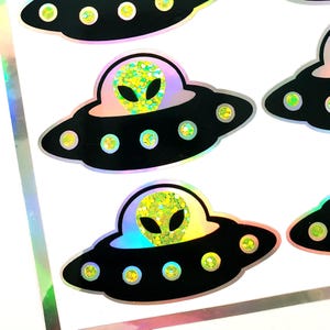 May include: A sheet of stickers featuring black UFOs with green glitter alien heads. The UFOs have yellow and green lights. The stickers have a holographic, iridescent border.