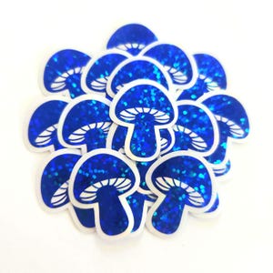 May include: A collection of blue mushroom stickers with a glittery, iridescent finish. Each sticker features a white outline and gills, creating a whimsical and decorative design. The stickers are arranged in a pile against a white background.