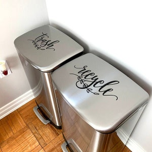 May include: Two silver trash cans with black labels. The label on the left can says "trash" and the label on the right can says "recycle".
