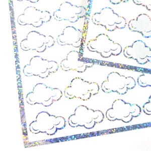 May include: Sheets of cloud-shaped stickers with a holographic, glittery border. The stickers are white with a cloud outline. The border is a shimmering silver color. The stickers are on a white background.