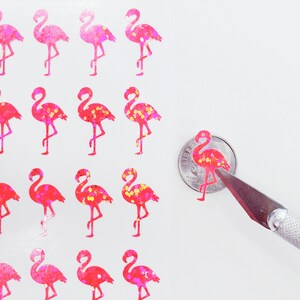 May include: Sheet of pink and red glitter flamingo stickers. The stickers vary in shades of pink and red, with some featuring yellow glitter accents. A small sticker is being cut with a craft knife.