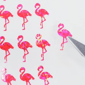 May include: Pink flamingo stickers with glitter accents are arranged on a white surface. A craft knife is positioned near one of the stickers. The stickers are in a repeating pattern, and the flamingo design is detailed.