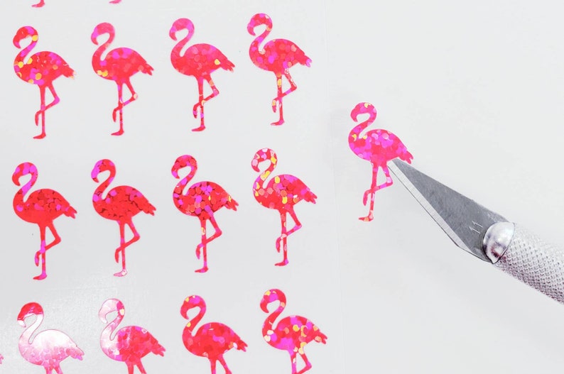 May include: Pink flamingo stickers with glitter accents are arranged on a white surface. A craft knife is positioned near one of the stickers. The stickers are in a repeating pattern, and the flamingo design is detailed.