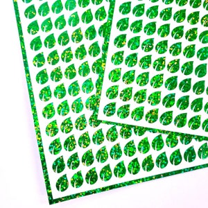 May include: Two sheets of green glitter paper with a repeating pattern of leaf shapes cut out. The glitter is iridescent and reflects light in different colors.
