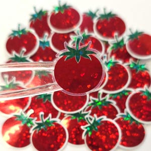 May include: A collection of red tomato-shaped embellishments with green stems and leaves. The tomatoes have a glittery, reflective surface. A clear plastic tool is holding one of the tomato embellishments.