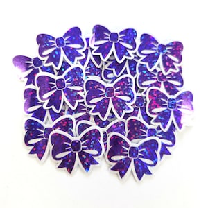 May include: A collection of purple bow-shaped embellishments with a glittery finish. Each bow has a white outline and a central knot detail. The bows are scattered on a white surface, creating a decorative effect. These are suitable for crafts.
