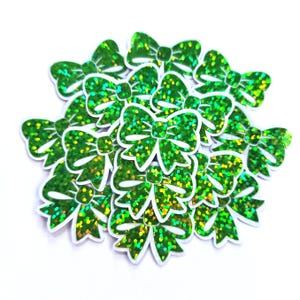 May include: A collection of green glitter bow-shaped embellishments with white outlines. The bows are clustered together, creating a festive and decorative arrangement. The glitter adds a sparkling effect to the bows.