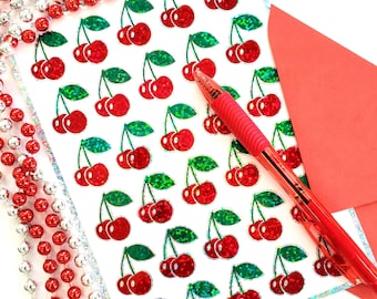 May include: A white card with a repeating pattern of red cherries with green stems and leaves. The cherries have a holographic finish. A red envelope and a silver pen are lying on top of the card.