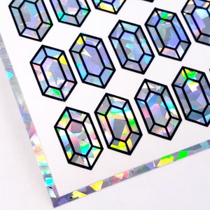 May include: Sheet of iridescent crystal-shaped stickers with black outlines. The crystals have a multifaceted design, reflecting a spectrum of colors. The stickers are arranged on a white background, with a matching iridescent border.