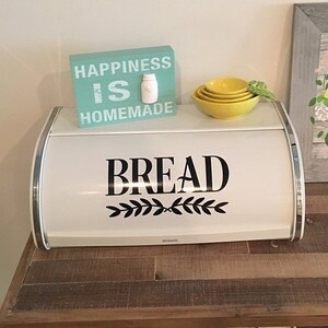 May include: A white metal bread box with a roll-top lid, featuring the black text BREAD and a leaf graphic. On top sits a teal wooden sign with the white text HAPPINESS IS HOMEMADE and a small jar icon, plus a stack of three yellow bowls.