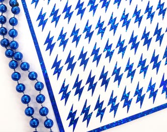 May include: A white rectangular sheet featuring a repeating pattern of blue lightning bolt shapes with a holographic finish, bordered by a blue holographic frame, accompanied by a strand of shiny blue spherical beads on the left side.