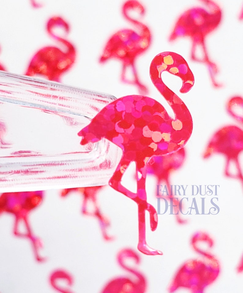 May include: Pink flamingo decal with glitter accents. The decal is a vibrant shade of pink with a textured, glittery surface. The flamingo is standing on one leg. The background features additional flamingo decals and the text "FAIRY DUST DECALS".