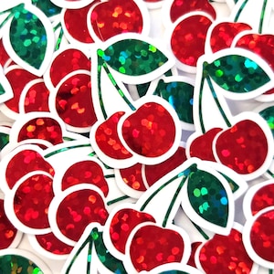 May include: A collection of cherry stickers featuring a vibrant red holographic finish on the fruit and a shimmering green holographic finish on the leaves. Each sticker has a distinct white border outlining the pair of cherries and the single leaf.