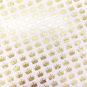 May include: A pattern of small, gold-colored crown stickers on a white background. The crowns have a textured, metallic appearance, and are evenly spaced across the surface. The image suggests a theme of royalty or celebration.