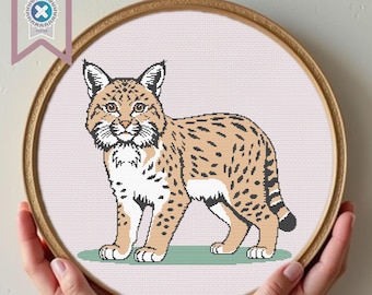 May include: A cross-stitch pattern of a bobcat, displayed in a wooden embroidery hoop. The bobcat is depicted in shades of tan, brown, and black, with a white chest and belly. The background is a light pink fabric. The hoop is held by hands.