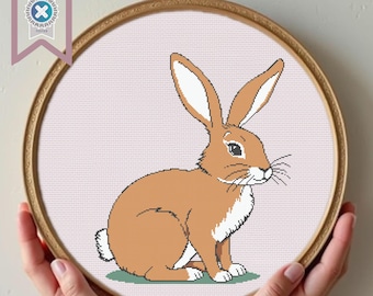 May include: A brown rabbit with white fur on its chest and a white tip on its tail sits on a green patch of grass. The rabbit is in a round embroidery hoop with a light pink background.