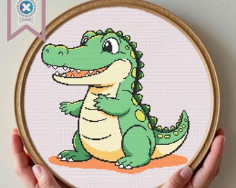 May include: A cross-stitch embroidery of a cartoon alligator in a wooden hoop. The alligator is green with yellow accents, a white belly, and an open mouth. The background is light pink. The hoop is held by hands.