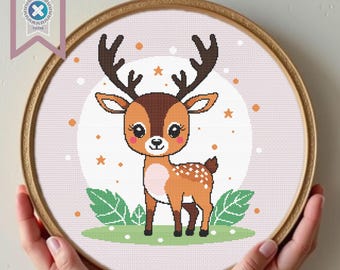 May include: A cross-stitch embroidery hoop featuring a cartoon deer with large eyes, antlers, and white spots. The deer stands on green grass with leaves, set against a pink background with stars and dots. The hoop is held by hands.