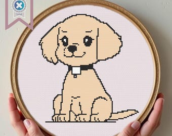Può includere: Un ricamo a punto croce di un cucciolo color camoscio chiaro con dettagli neri, con un collare nero. Il cucciolo è centrato in un cerchio di legno rotondo, tenuto in mano. Lo sfondo è un tessuto rosa chiaro. Un banner di download PDF si trova nell'angolo in alto a sinistra.