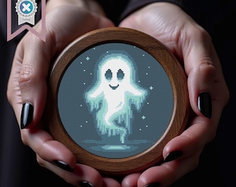 Può includere: Un disegno a punto croce di un fantasma amichevole in un telaio da ricamo in legno. Il fantasma è bianco con un volto sorridente e una forma fluida ed eterea, su uno sfondo blu scuro con piccole stelle bianche. Il telaio di legno è tenuto in mano.