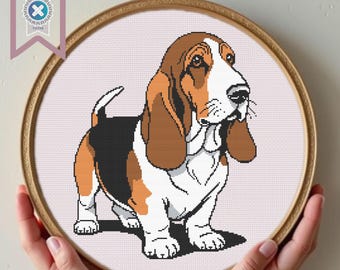 Può includere: Schema a punto croce di un cane Basset Hound in un telaio da ricamo in legno. Il cane è raffigurato in tonalità marroni, nere e bianche, con orecchie lunghe e un muso cadente. Lo sfondo è in tessuto rosa chiaro. Badge "PDF Download" in alto a sinistra.