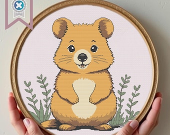 May include: A cross-stitch pattern featuring a cute cartoon quokka with brown fur, big eyes, and a small nose. The quokka is standing on a pink background with green leaves.