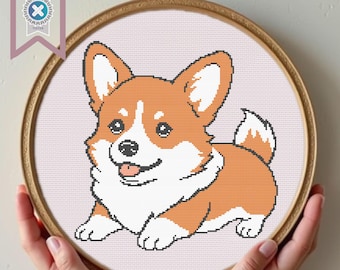 May include: A cross-stitch embroidery of a smiling Corgi dog in a round wooden hoop. The dog is orange, white, and black, with its tongue sticking out. The background is light pink. A banner in the upper left corner reads "PDF Download".