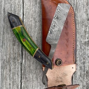 May include: A handcrafted knife with a green and black handle, gold accents, and a patterned blade, partially sheathed in a brown leather case. The knife is open, revealing the blade's intricate design.