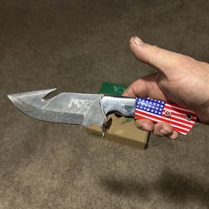 May include: A Damascus steel knife with a hook-shaped tip. The handle features a red, white, and blue American flag design. The blade has a unique pattern and the knife is held in a hand.