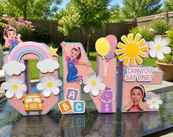 May include: A colorful, decorative "ONE" sign with a rainbow, flowers, and a school bus. The letters are adorned with images of a girl, balloons, a sun, and the text "CAN YOU SAY ONE?"