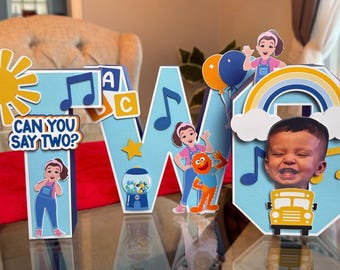 May include: A colorful birthday decoration spelling out "W" with a sun, musical notes, and a rainbow. The decoration includes cartoon characters, a gumball machine, and a school bus. The text "Can You Say Two?" is also present.
