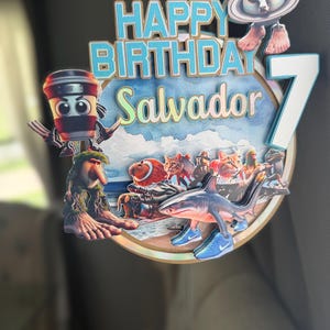 Może przedstawiać: Kolorowa dekoracja na tort urodzinowy z napisem "HAPPY BIRTHDAY Salvador" w kolorze niebieskim i białym. Wyświetlana jest liczba 7. Dekoracja zawiera postacie z kresk&oacute;wek, w tym rekina w trampkach, filiżankę kawy i kosmitę.