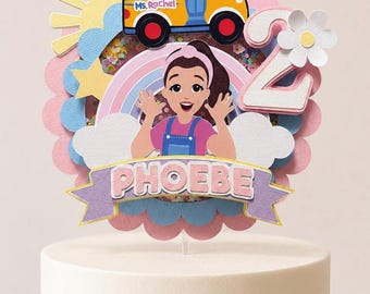 May include: A layered cake topper featuring a Ms. Rachel illustration, a yellow school bus with the text Ms. Rachel, a rainbow, clouds, a sun, a star, a white flower, and the number 2. A purple banner at the bottom displays the name PHOEBE.