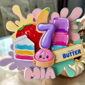 May include: A layered cake topper featuring a purple number 7, a red strawberry, a yellow banana, a slice of rainbow cake, a pink dumpling, purple and blue Nee Doh blocks, and a stick of salted butter, with the name MIA at the bottom.