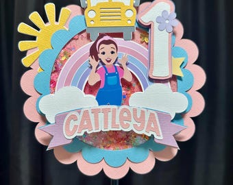 May include: A vibrant birthday cake topper with a yellow school bus, a cartoon girl, a rainbow, and the number 1. The name "CATTLEYA" is displayed on a banner. The topper is decorated with pink, blue, and yellow accents.
