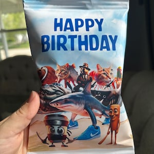 May include: A white and blue Happy Birthday snack bag featuring a cartoon shark, a cat, and a stick figure on a beach.